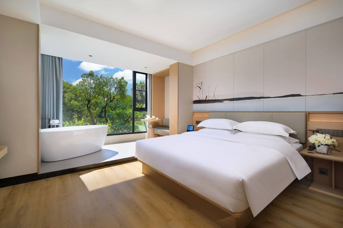 A modern bedroom with a large bed, a bathtub, and a view of greenery through a large window.