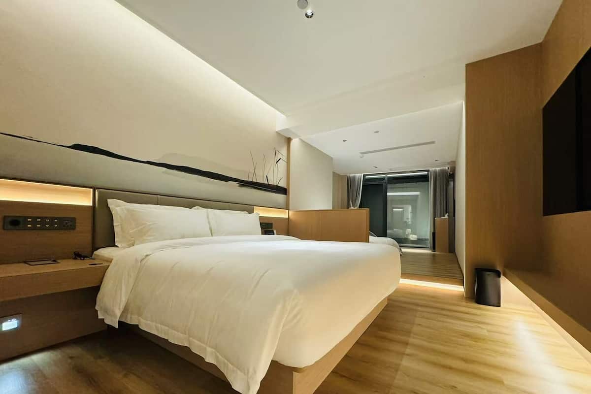 A modern hotel room with a large bed, bedside controls, a wooden floor, and a view of the hallway.