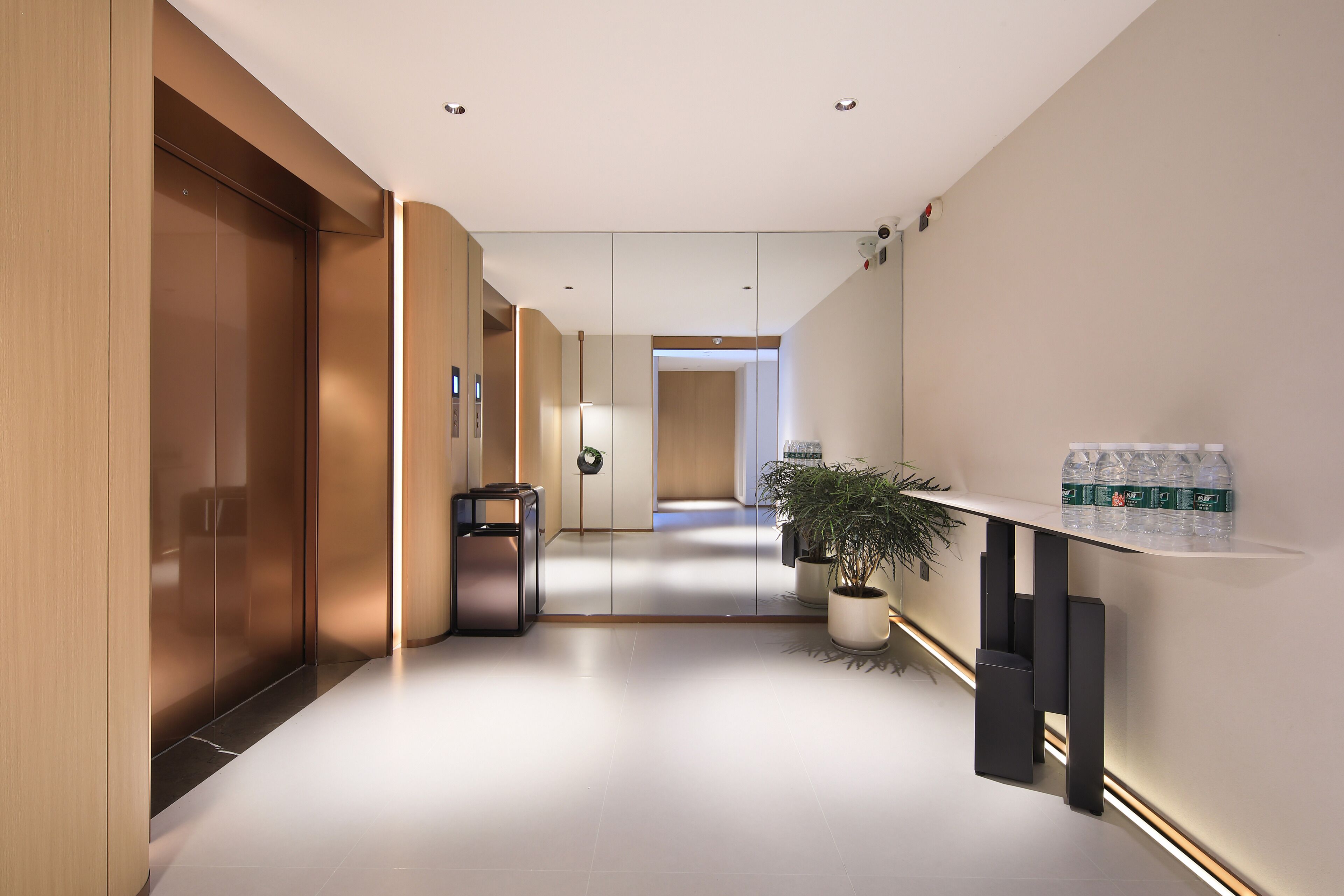 A modern hotel hallway with a wooden elevator, a trash bin, a potted plant, and a shelf with bottled water.