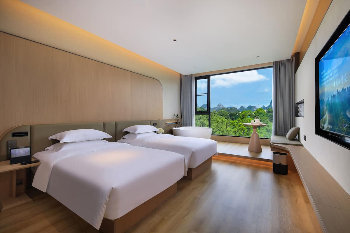 A modern hotel room with two beds, a desk, a TV, and a large window offering a view of the outdoors.