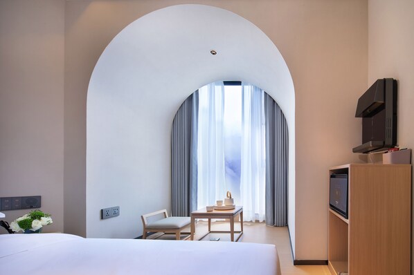 A hotel room with a large arched window, a bed, a desk with a chair, a television, and a small table with a vase of flowers. - GuiLin Lijiang Wangyue Hotel (Guilin)