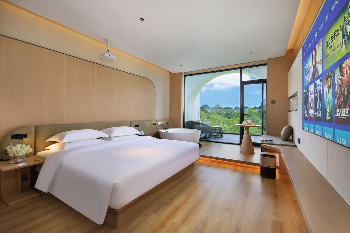 A modern hotel room with a large bed, bedside tables, a small seating area, and a large window offering a view of the outdoors.