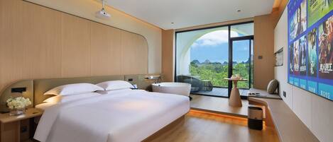 A modern hotel room with a large bed, bedside tables, a small seating area, and a large window offering a view of the outdoors.