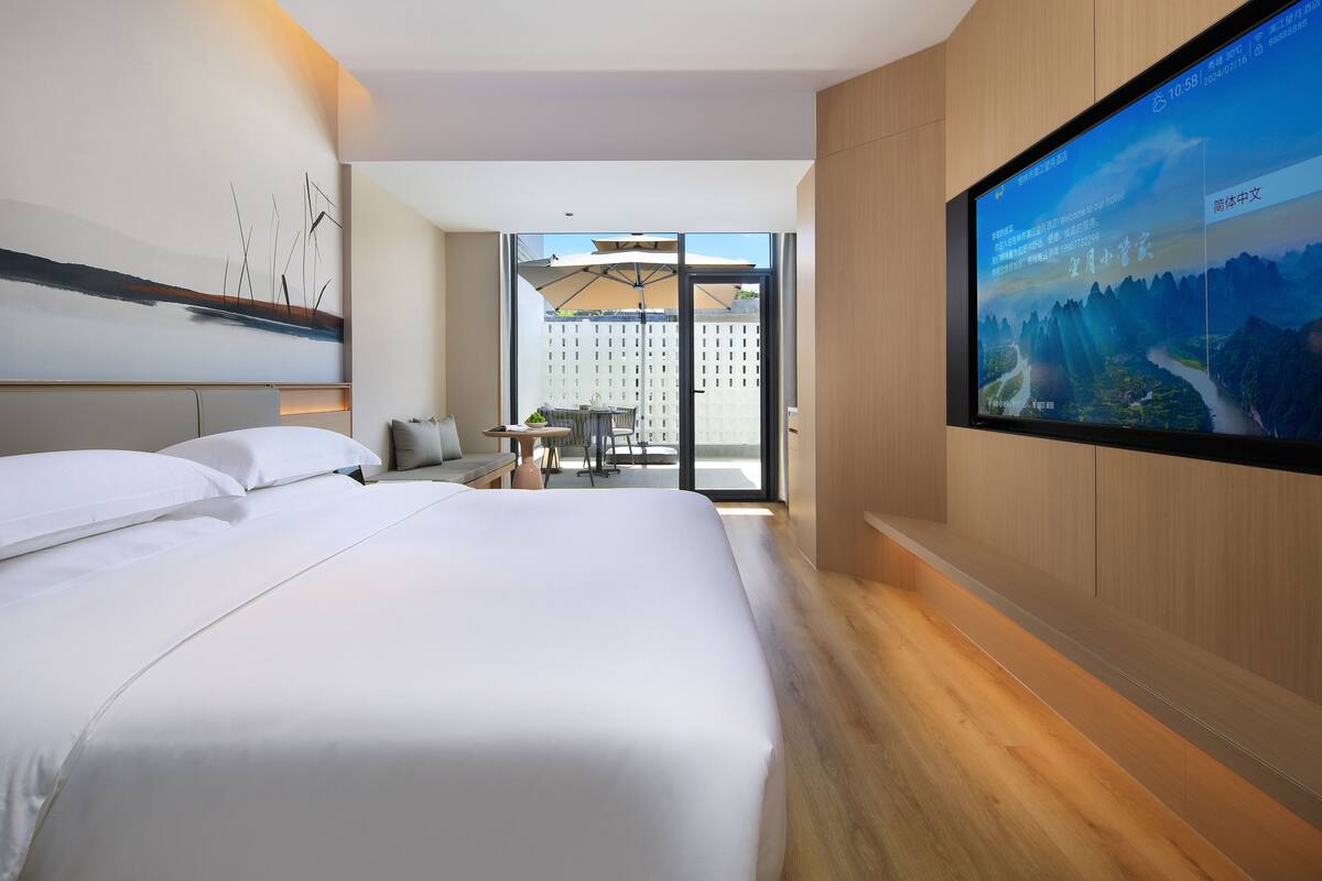 A modern hotel room with a large bed, a TV on the wall, and a balcony with outdoor seating.