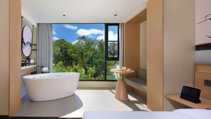 Une chambre d’hôtel moderne dotée d’une grande baignoire, d’une table de chevet avec une tablette et offrant une vue sur une végétation luxuriante grâce à de grandes fenêtres.
