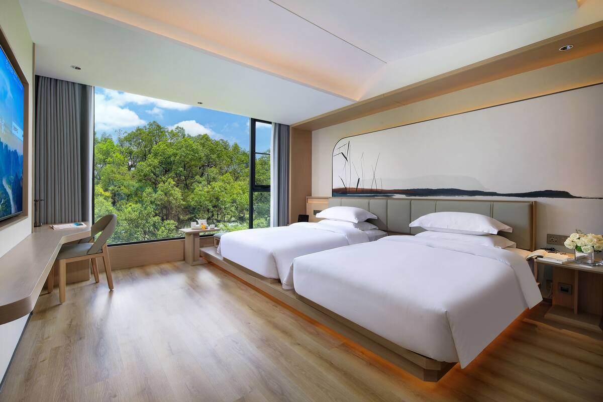A modern hotel room with two beds, a desk, and a large window offering a view of trees.