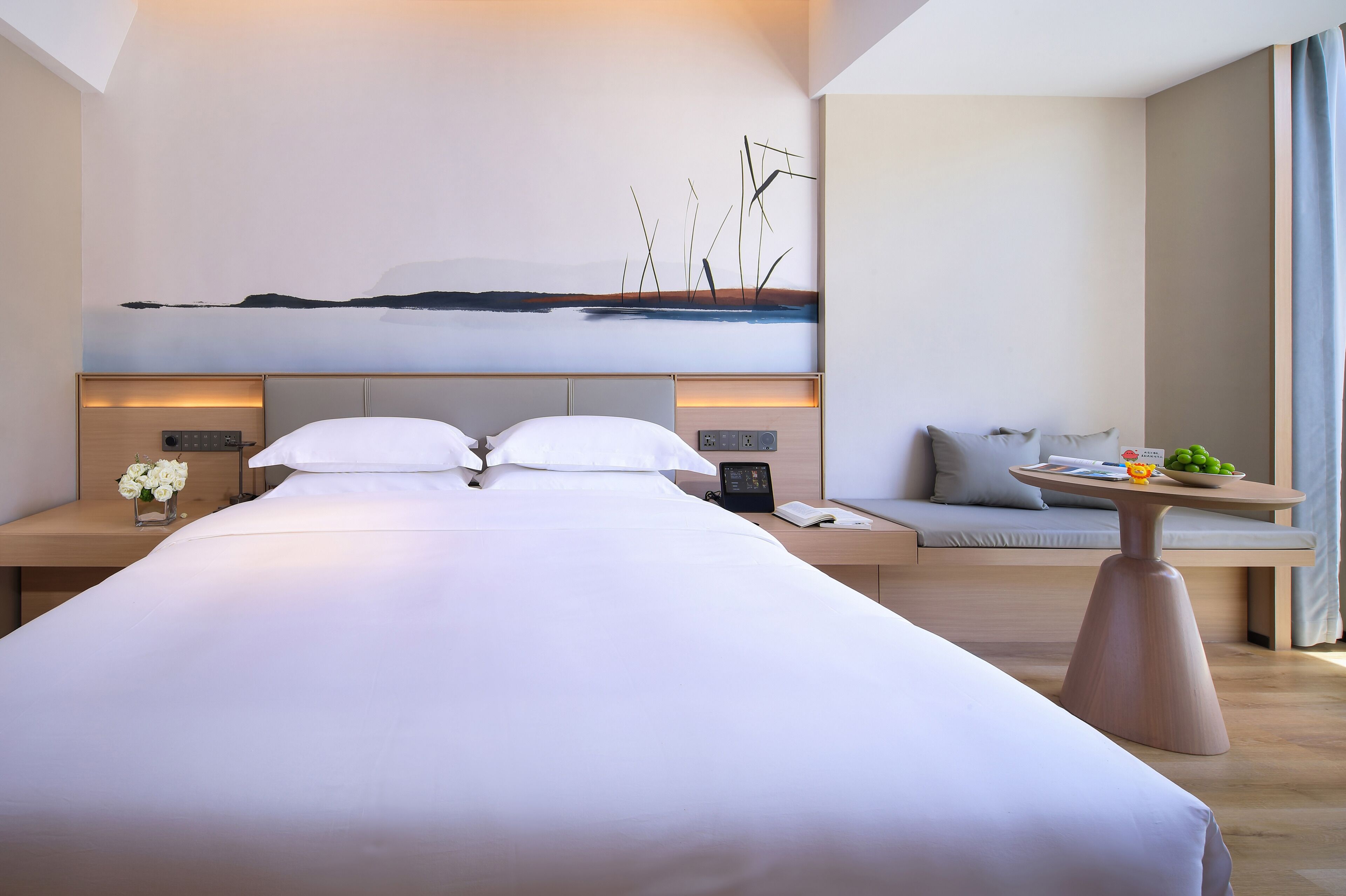 Une chambre d’hôtel moderne avec un grand lit, des tables de chevet, une petite table avec des fruits, un canapé et une œuvre d’art accrochée au mur.