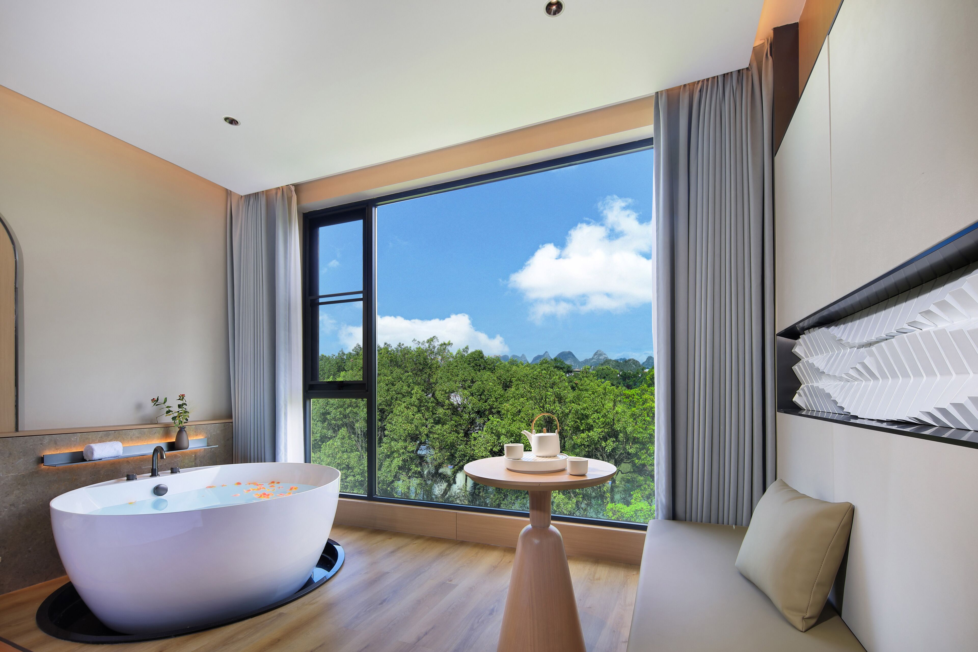 A modern bathroom with a freestanding bathtub, a round table with a teapot and cups, and a large window offering a view of a forested landscape.