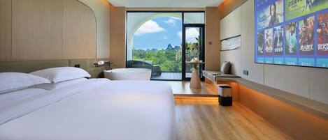 A hotel room with two beds, a large window with a view of greenery, and a wall-mounted screen displaying movie posters.