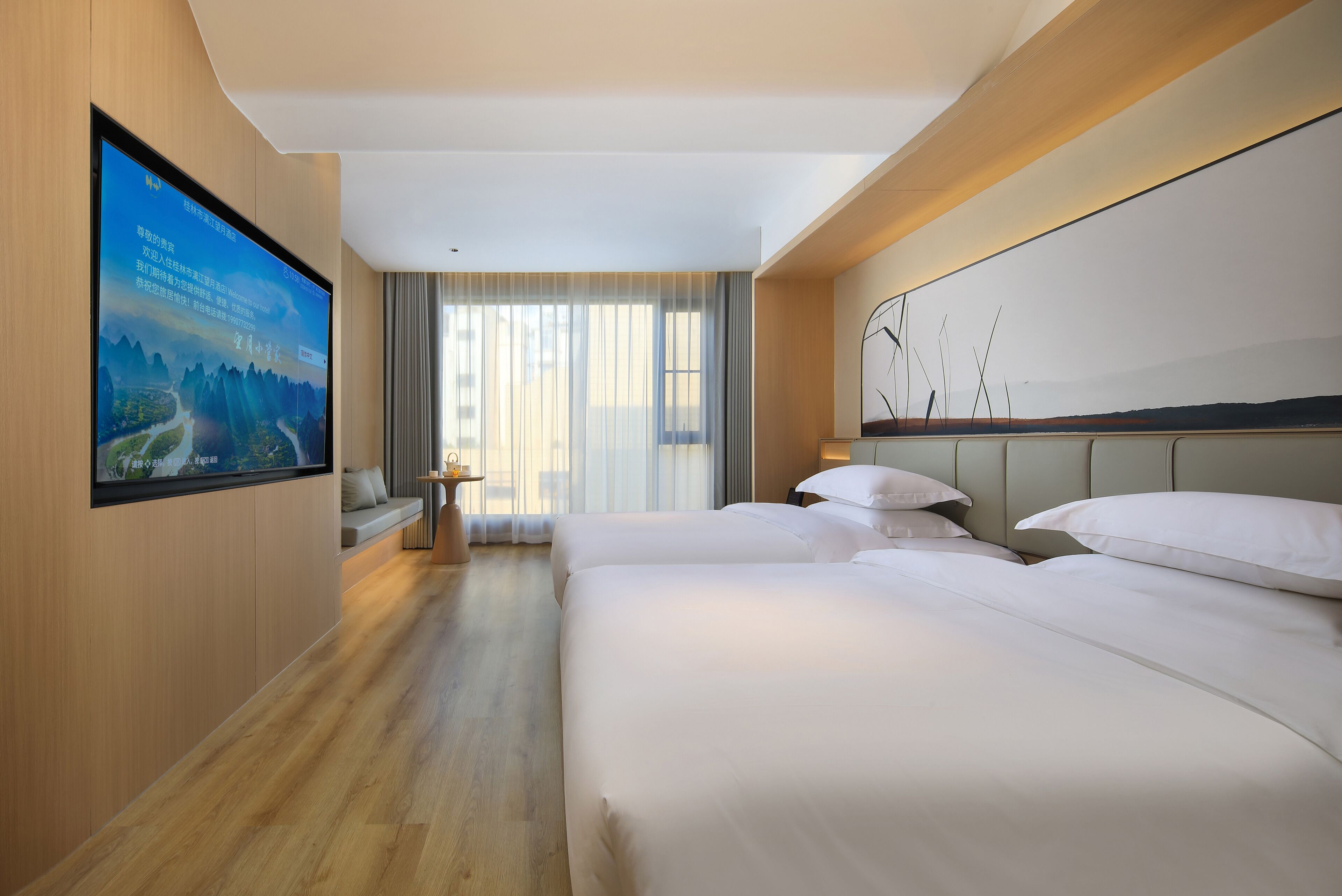 A hotel room with a large flat-screen TV mounted on the wall, a bed with white linens, a bedside table with a lamp, and a view of the outdoors through large windows.