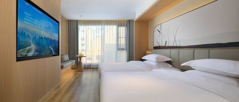 A hotel room with a large flat-screen TV mounted on the wall, a bed with white linens, a bedside table with a lamp, and a view of the outdoors through large windows.