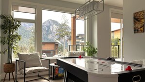 A modern living room with a foosball table, large windows offering mountain views, and a hanging light fixture.
