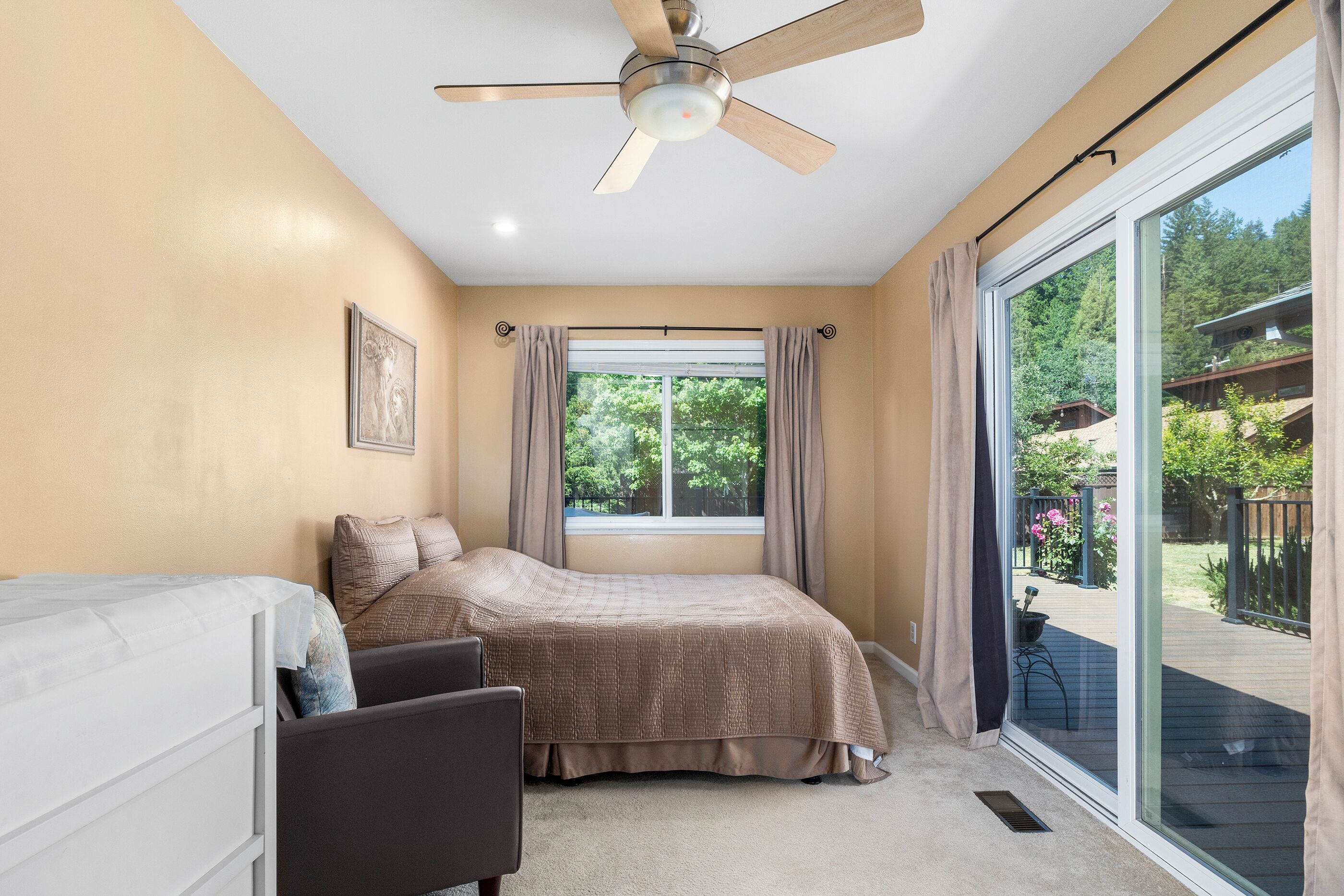 A bedroom with a bed, a chair, a ceiling fan, a window with curtains, and a view of the outdoors.