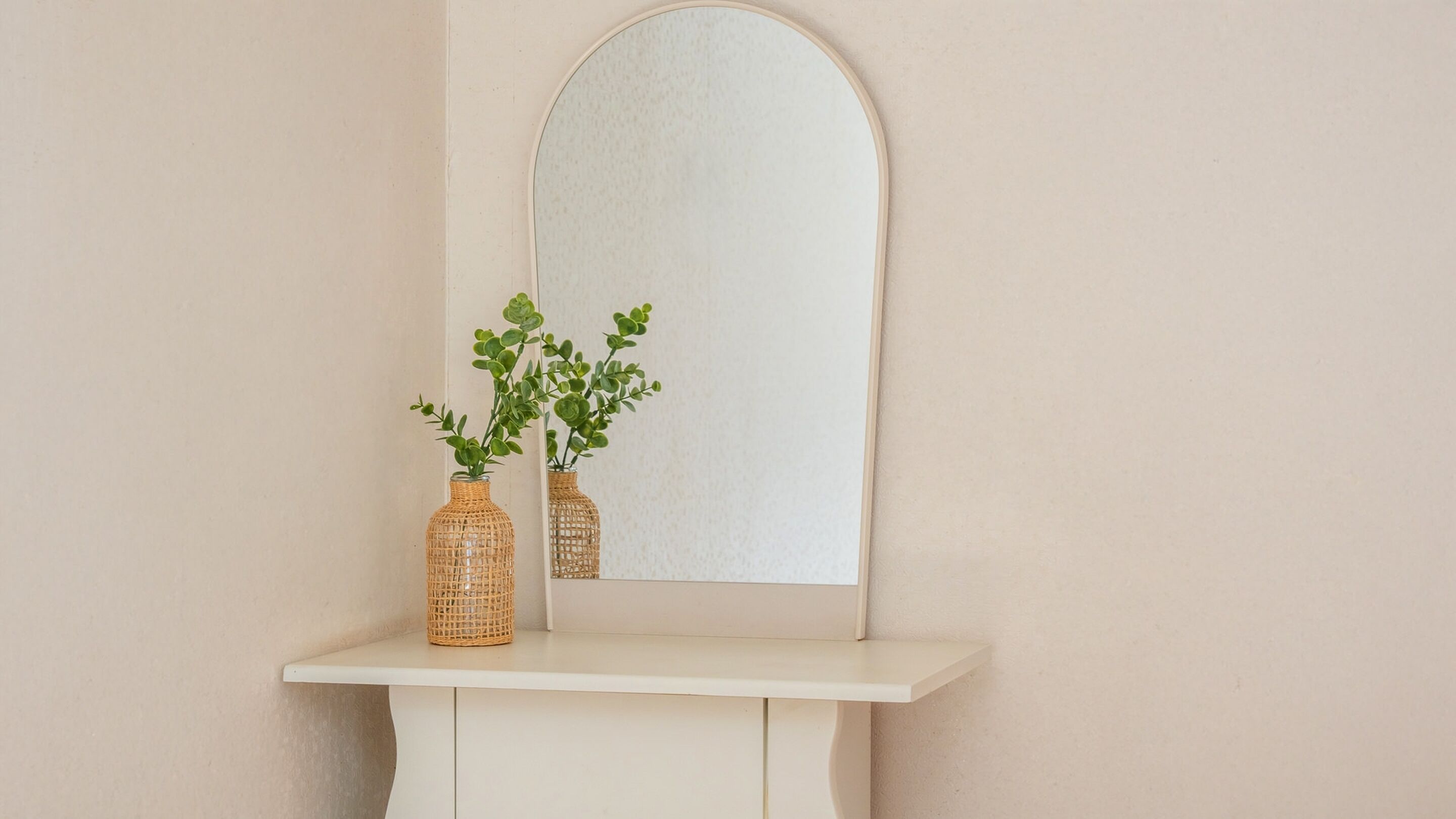 Une coiffeuse blanche, ornée de deux vases en osier contenant des plantes vertes, est placée devant un miroir à cadre blanc.