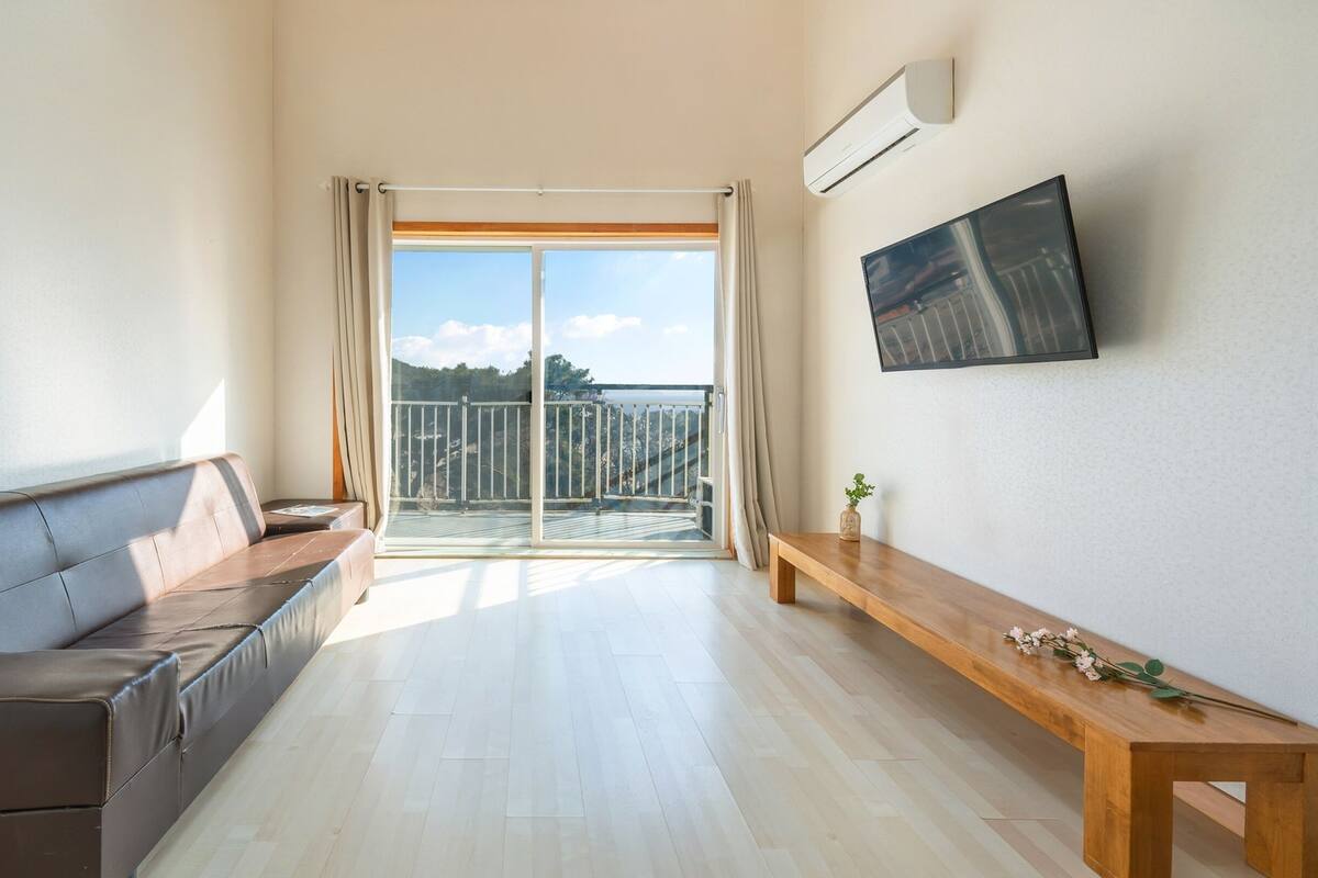 Un salon moderne avec un canapé en cuir, un banc en bois et une grande fenêtre donnant sur un balcon et des arbres.