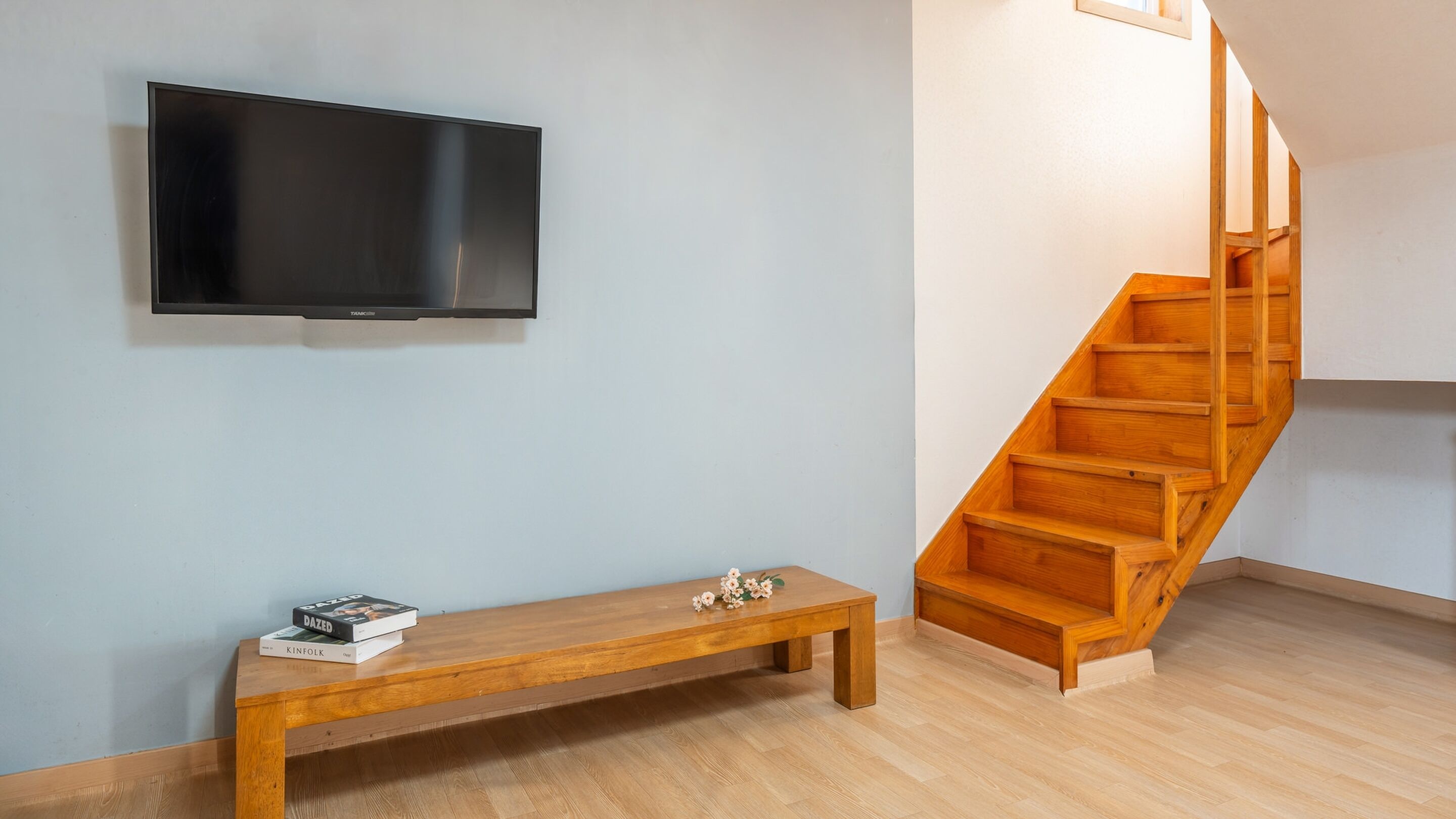 A modern living room with a wooden bench, a flat-screen TV mounted on the wall, and a wooden staircase.