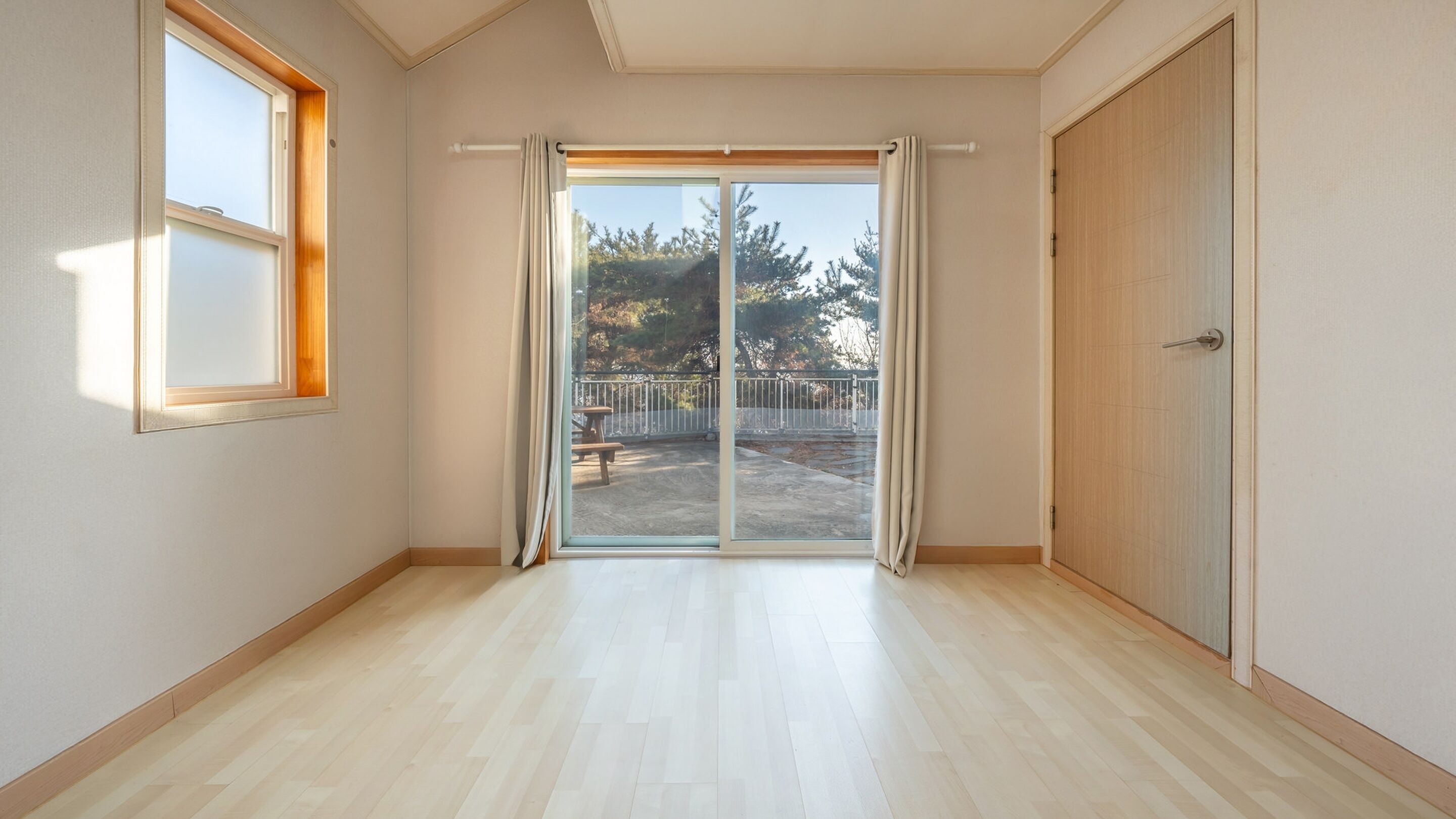 A room with a sliding glass door leading to an outdoor area, a wooden floor, and a wooden door.