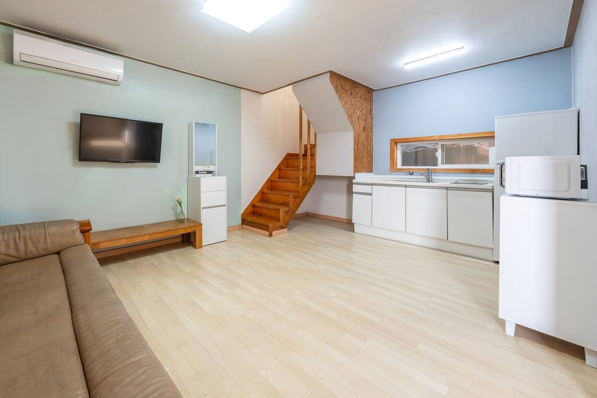Un salon moderne comprenant un canapé, un téléviseur à écran plat, un banc en bois et une cuisine avec des meubles blancs et un four à micro-ondes.