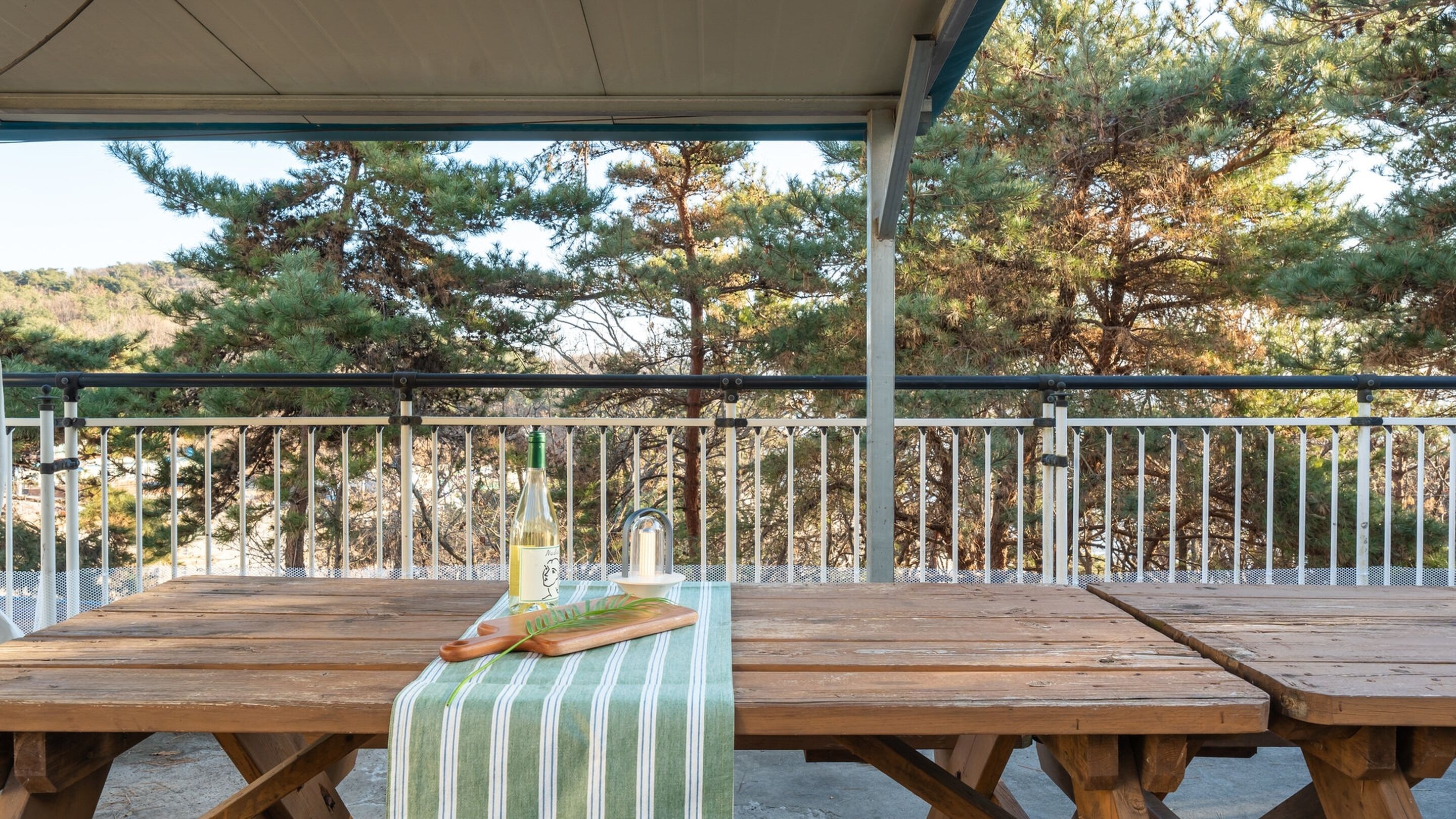 An outdoor wooden table with a green and white striped tablecloth, a bottle of wine, and a candle on a covered patio with a metal railing and trees in the background.