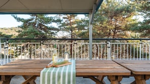 Ein Holztisch im Freien mit einer grün-weiß gestreiften Tischdecke, einer Weinflasche und einer Kerze auf einer überdachten Terrasse mit Metallgeländer und Bäumen im Hintergrund.