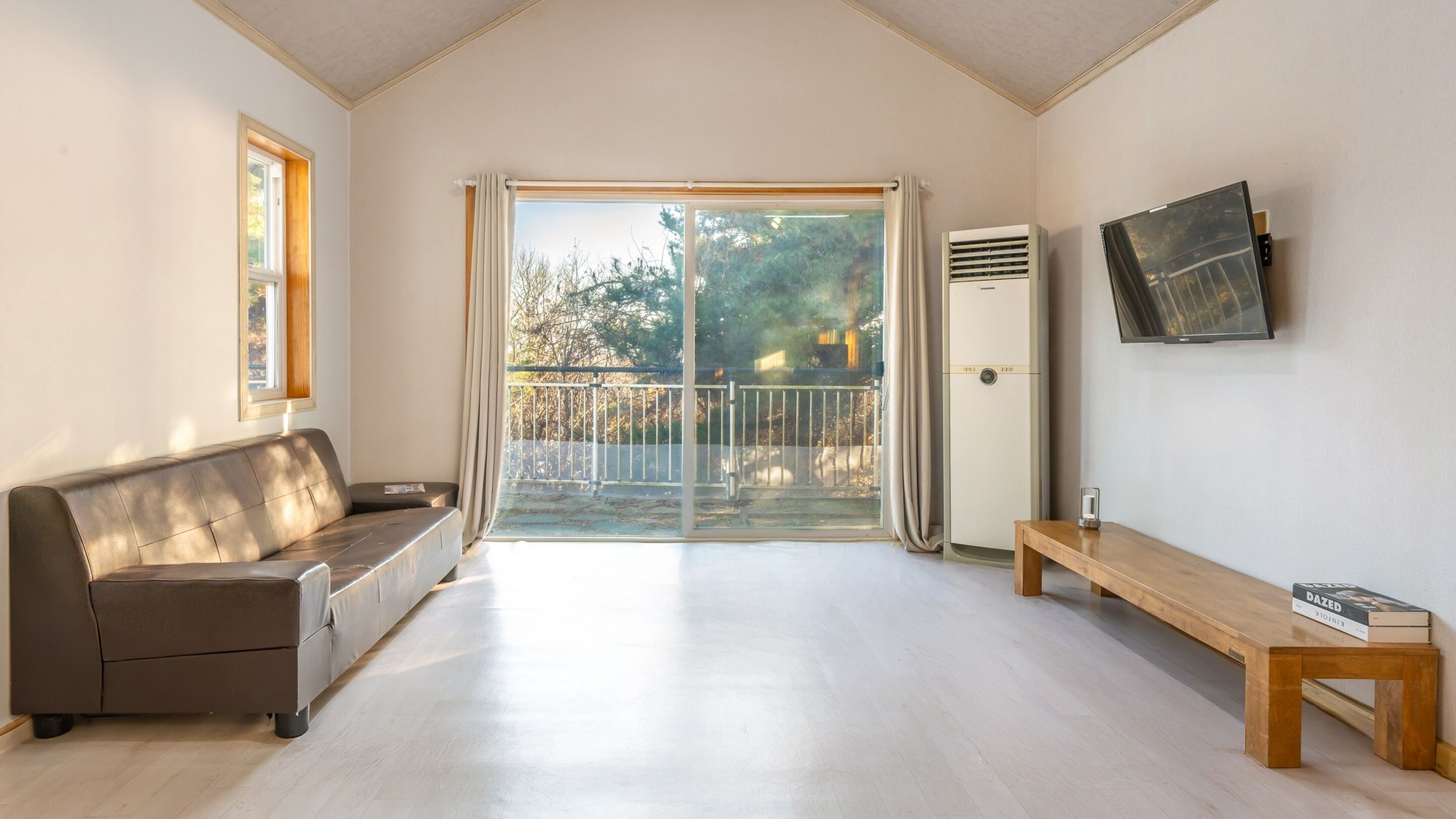 A spacious living room with a sofa, a wooden bench, a flat-screen TV, and a sliding glass door leading to an outdoor balcony.