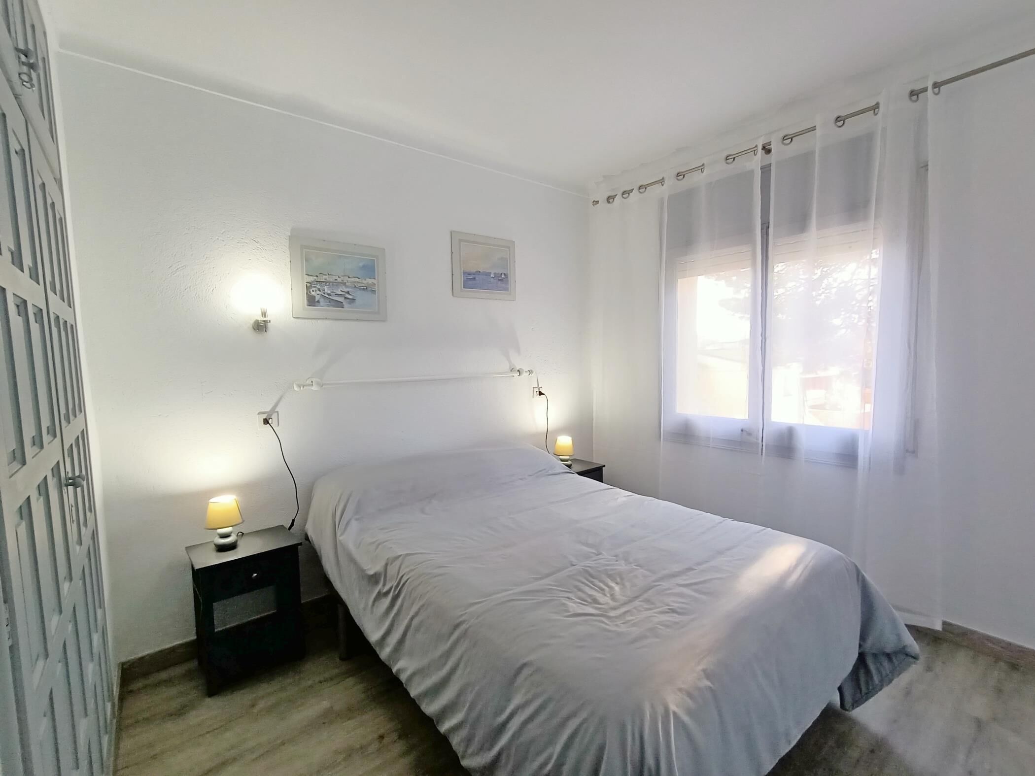 Ein Schlafzimmer mit einem Bett, zwei Nachttischen mit Lampen, einem Fenster mit Vorhängen und zwei Wandbildern.