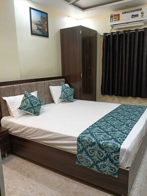 A neatly made bed with a patterned bedspread, a dark wooden wardrobe, and a framed picture on the wall. - JS Banaras Residency (Varanasi)