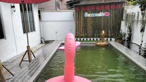 Een roze opblaasflamingo in een zwembadje, met een bamboestandaard en een rode parasol in de buurt.