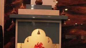 Une statue représentant une divinité éléphant, tenant une cloche, entourée de fleurs et d’une petite lampe posée sur une étagère.