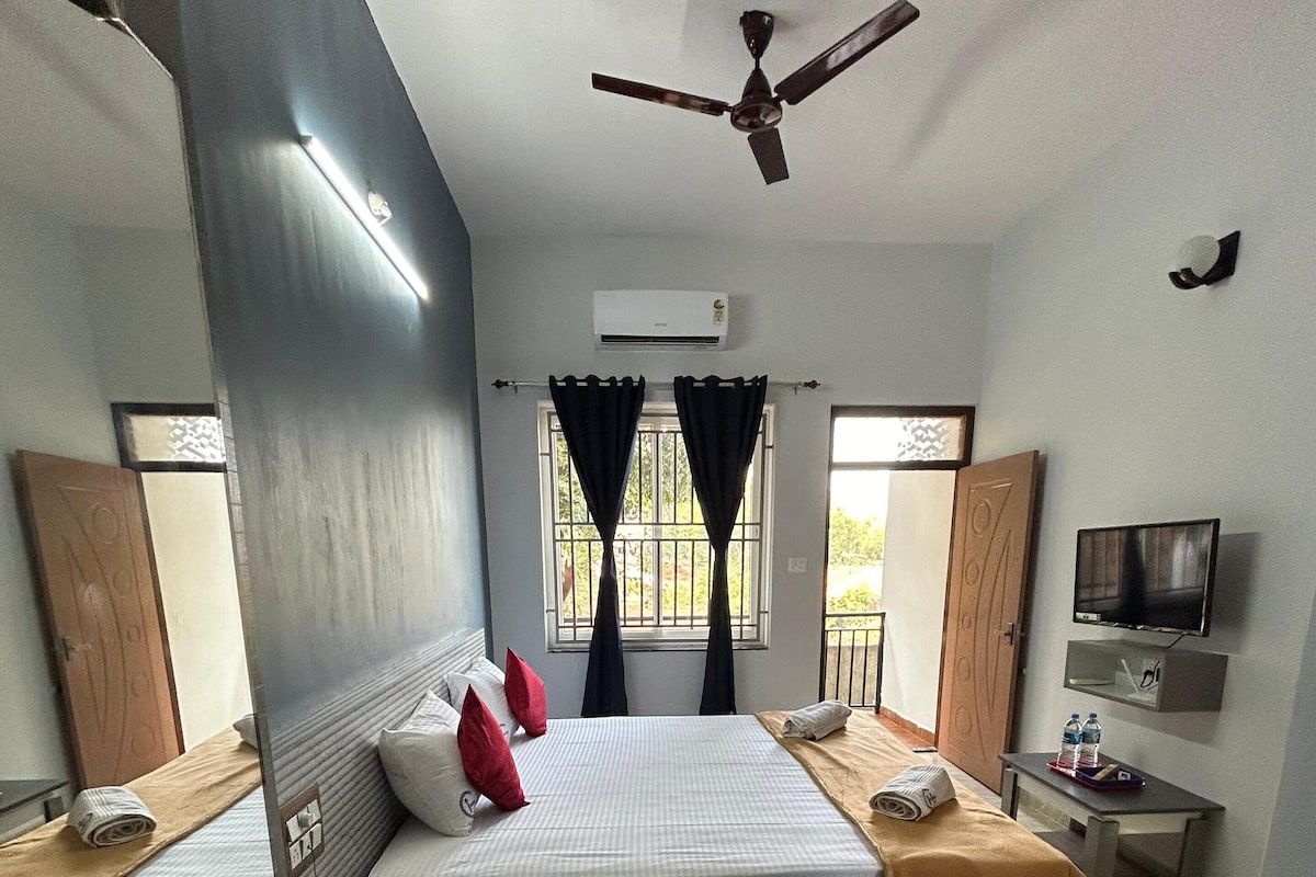 Una camera d'albergo con un letto grande, due comodini, una ventola a soffitto, un condizionatore d'aria, una televisione e una finestra con tende.