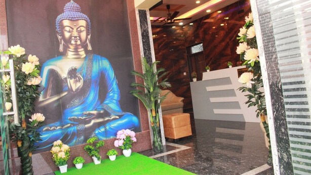 Une statue de Bouddha, vêtue d’une robe bleue, est placée sous une enseigne où l’on peut lire : « GOLDEN IB’S RESIDENCY ». Au premier plan, on aperçoit des plantes en pot et un comptoir d’accueil.