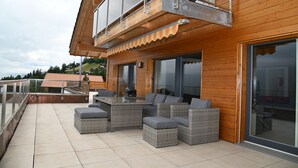 Un balcon en bois avec une porte vitrée, un espace salon extérieur et une vue sur les environs.