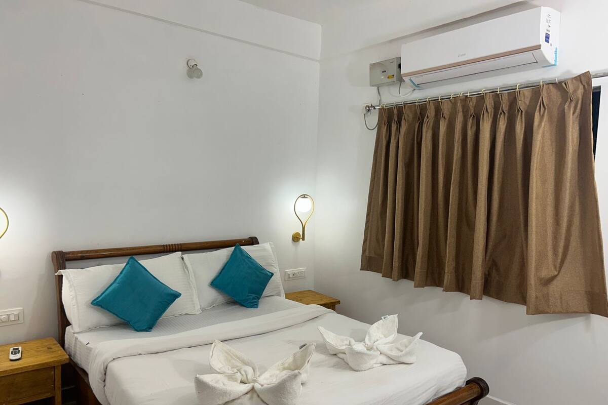 Uma cama bem arrumada com almofadas brancas e azul-turquesa, uma cabeceira de madeira e uma mesa de cabeceira branca com um pequeno objeto em cima.