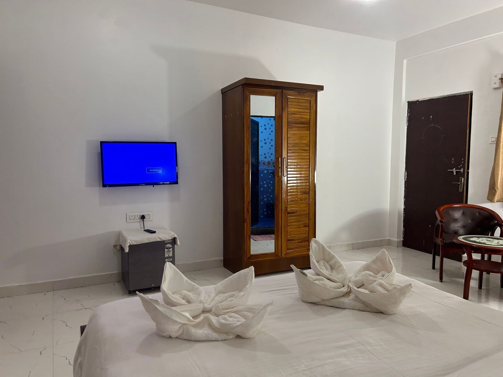 A hotel room with a bed, a TV mounted on the wall, a wardrobe, a desk with a chair, and a door with a curtain.