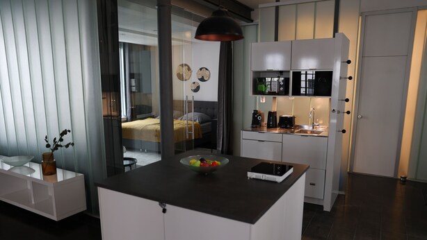 Et moderne køkken med lyse skabe, en central ø med frugt i en skål, og et spiseområde, der kan ses gennem en glasvæg.
