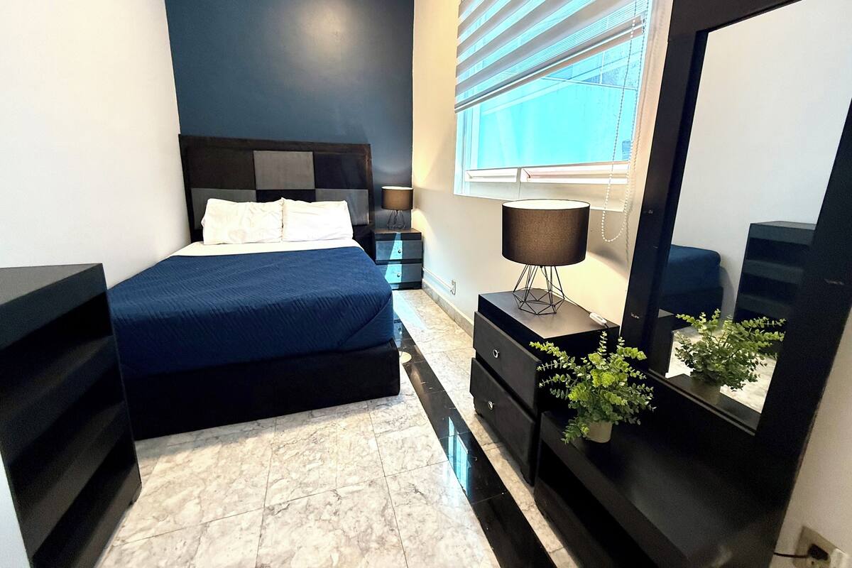 A bedroom with a bed, a nightstand, a lamp, a dresser, a mirror, and a potted plant.
