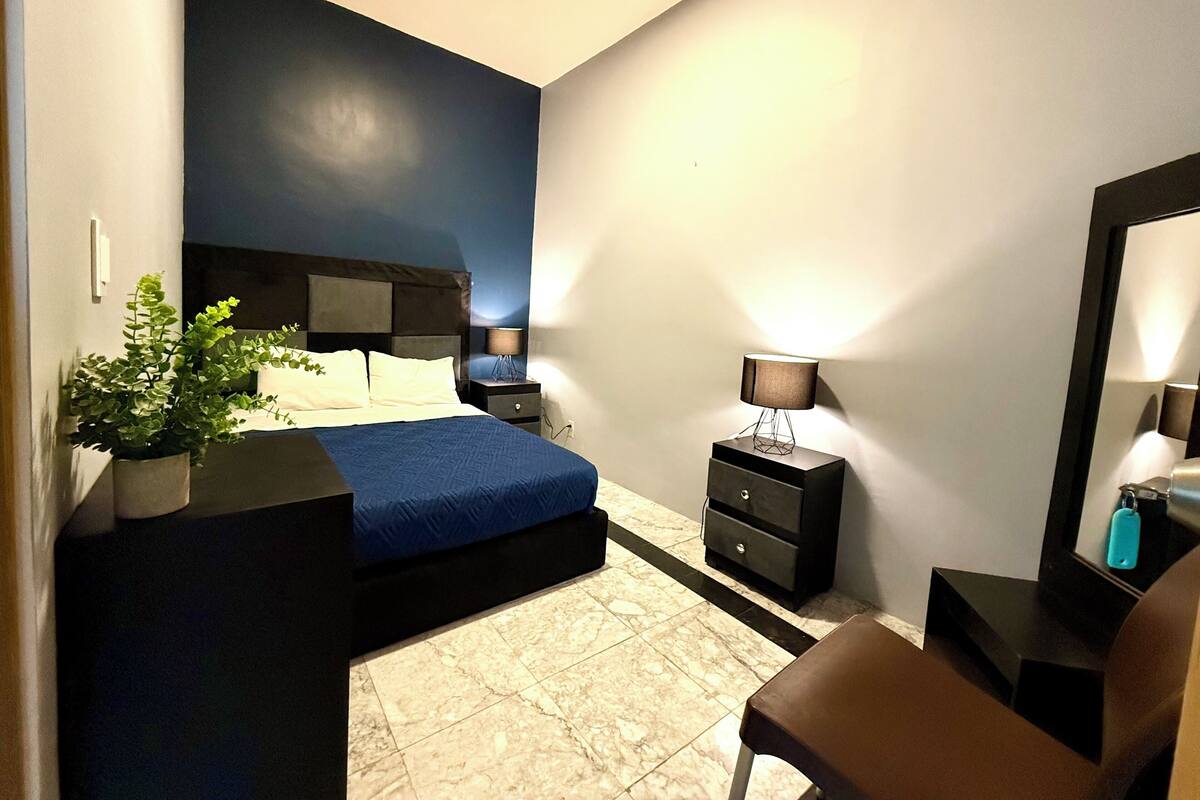 A bedroom with a bed, bedside tables, a desk, a chair, and a potted plant.