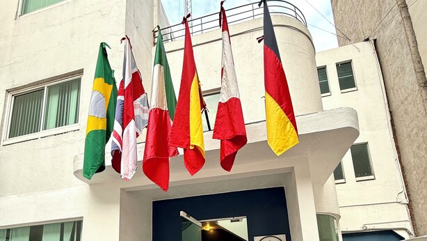 A building entrance with multiple flags hanging above a staircase leading to a room with a checkered floor. - ROOMIES HOSTEL Reforma - La Diana (Mexico City)