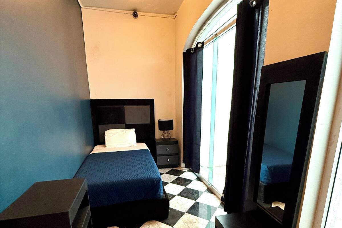 A hotel room with a checkered floor, a bed with a blue bedspread, a small table, a TV, and a window with curtains.