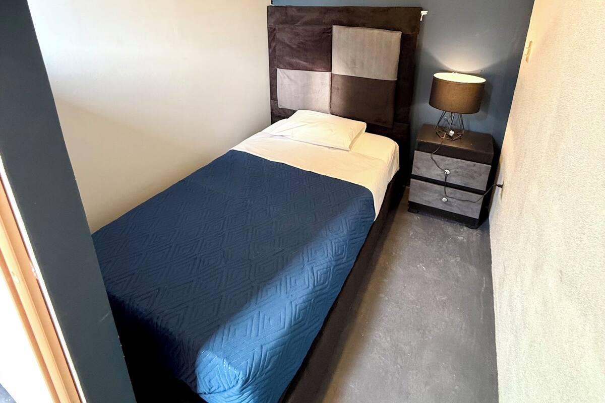 A small, compact bedroom with a single bed, a nightstand, and a lamp.