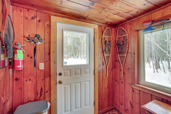A wooden cabin interior with a white door, a window with a view of trees, and a bench. - Fish, Hike & Relax: Riverfront Manistique Cabin (Manistique)