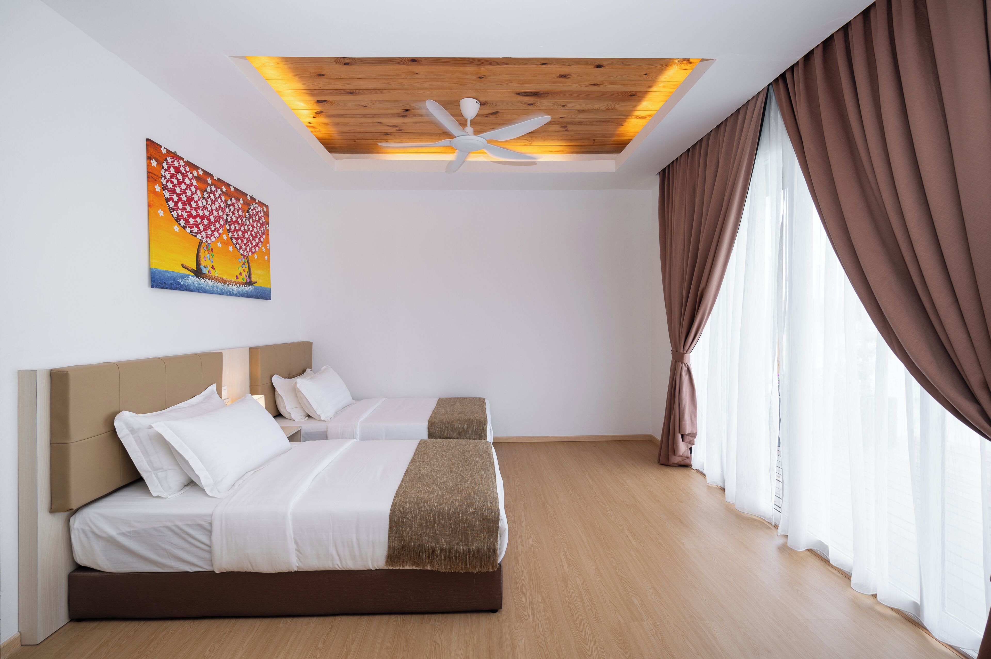 A bedroom with two beds, a ceiling fan, a painting, and large windows with curtains.