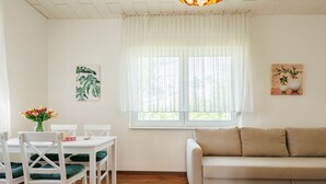 A dining area with a white table and chairs, a beige sofa, and a window with sheer curtains. - Green House by Interhome (Balatonmariafurdo)