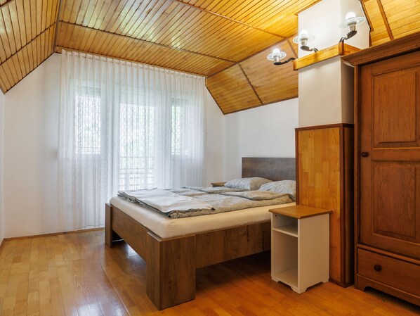 Un dormitorio con techo inclinado, una cama de madera, una mesita de noche, un armario y una ventana con cortinas traslúcidas.