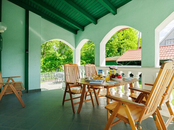 A covered patio with wooden furniture, a table set with breakfast items, and a view of a red-roofed building through arched windows.
