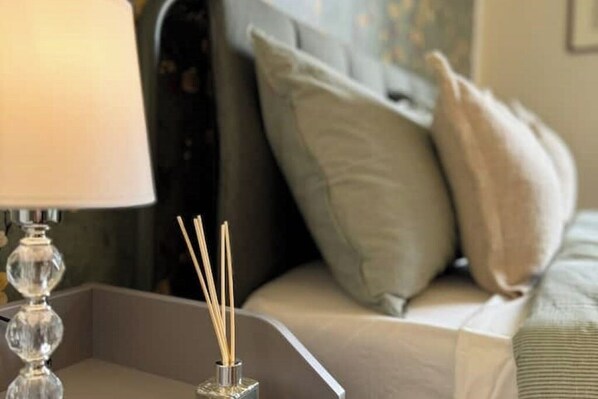 En seng med gråt sengetøj og to puder, et natbord med lampe og en rør diffusor.