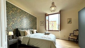 Een slaapkamer met een bed, nachtkastje, stoel en een raam met uitzicht op gebouwen.