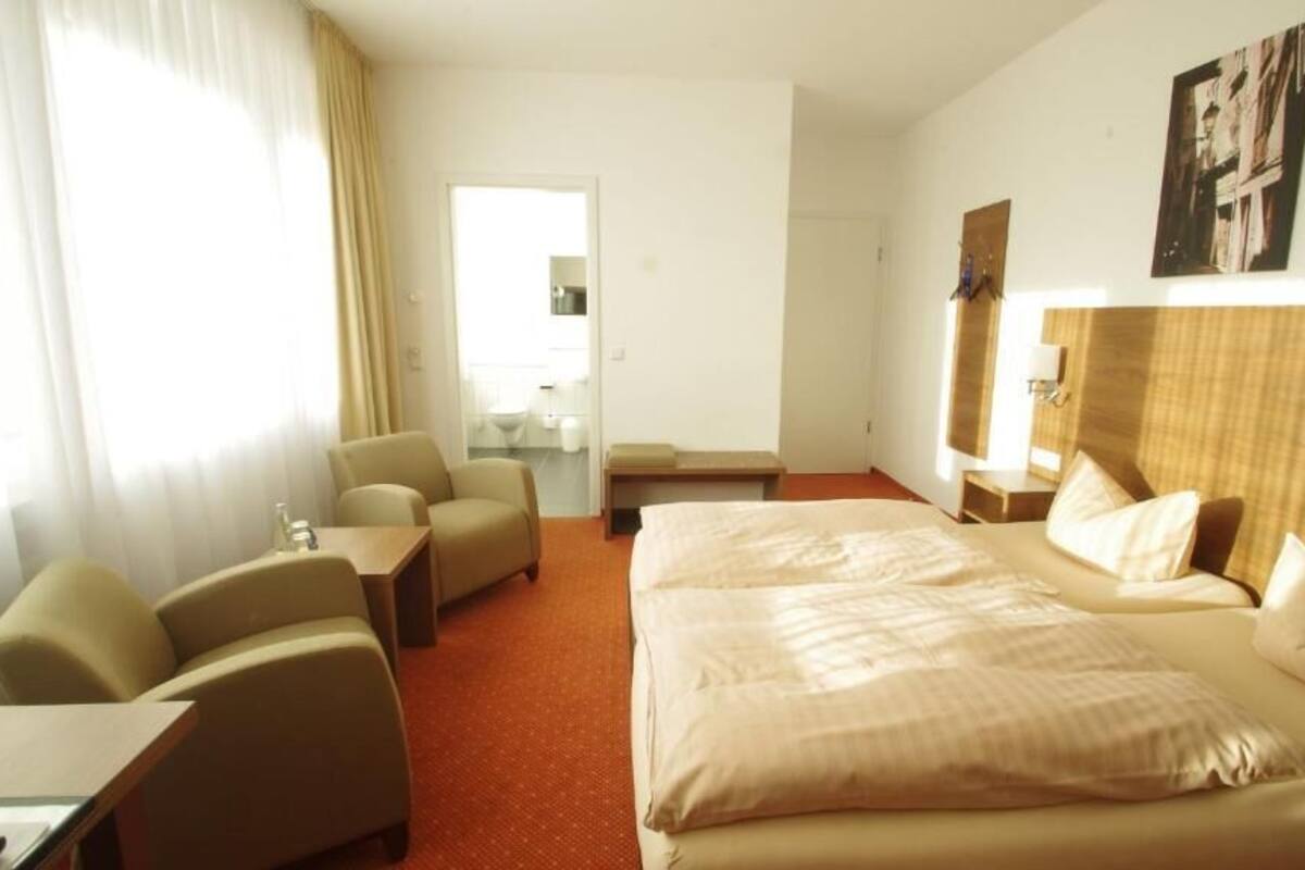 Habitación de hotel con una cama grande, dos sillones, una mesita y un escritorio. Se ve una ventana con cortinas, una puerta y una lámpara fijada a la pared.