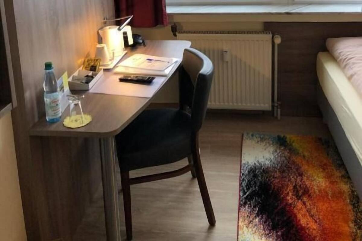 Una habitación de hotel con una cama, un escritorio con una silla, un televisor montado en la pared, una ventana que ofrece vistas a edificios y un radiador debajo de la ventana.