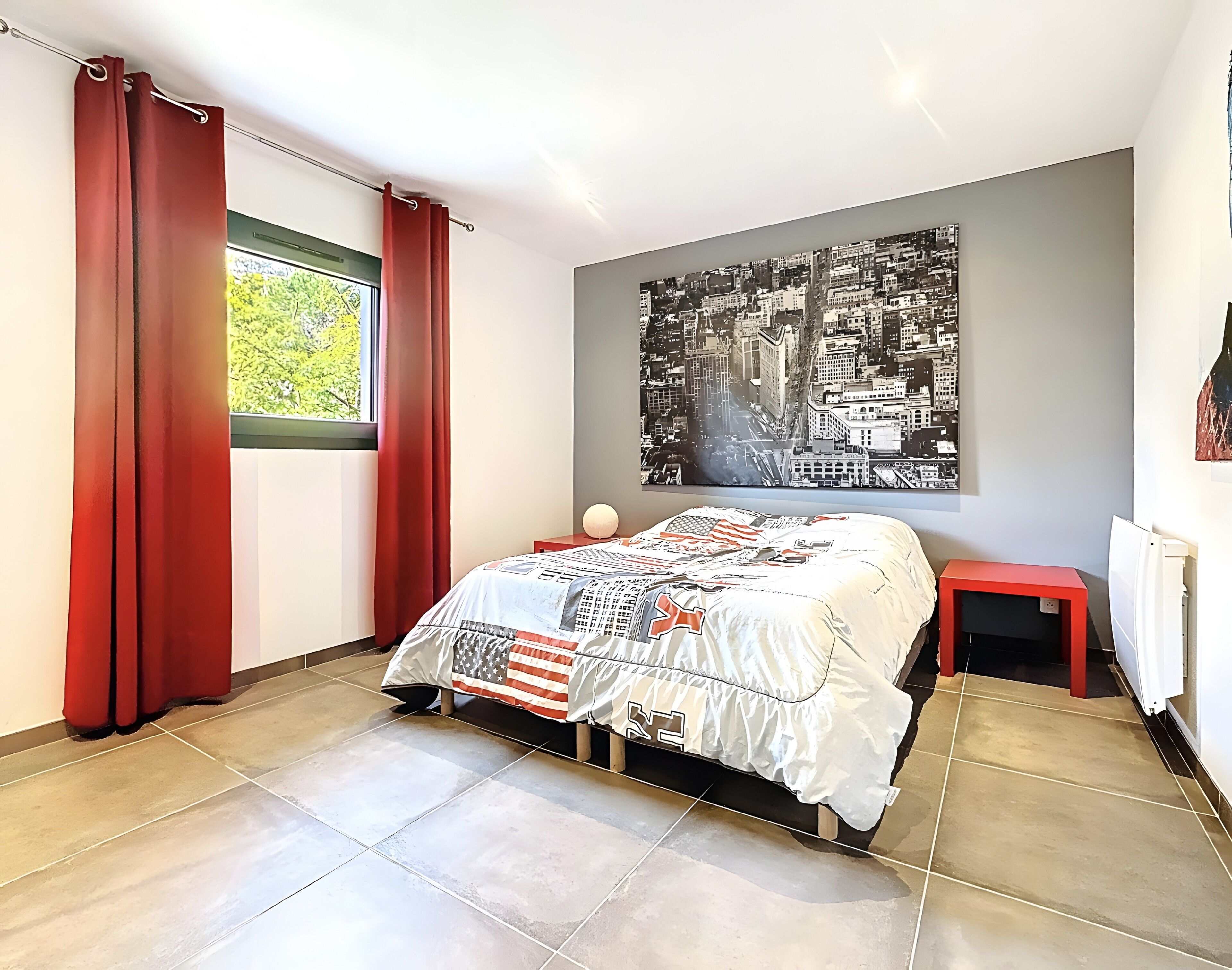 Ein Schlafzimmer mit Bett, Nachttisch, Fenster mit Vorhängen, einer Wandkunst mit Stadtansicht und einem Heizkörper.